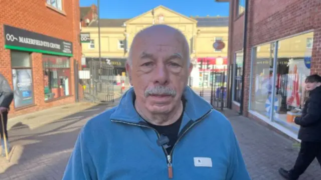 Aman wears a light blue quarter-zip top in a busy town centre. He has a grey moustache.