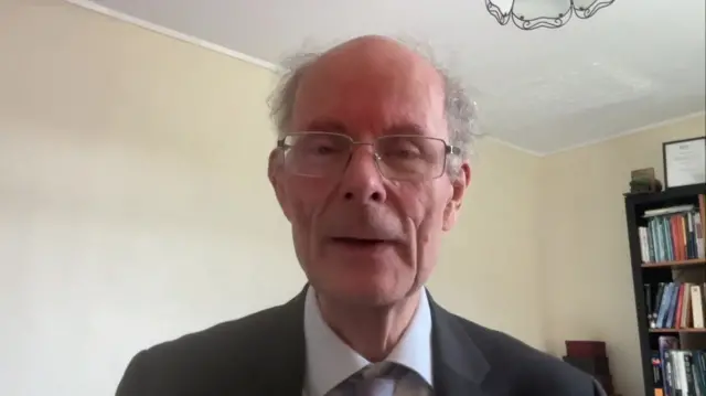 John Curtice ears a dark grey suit with a light grey shirt and tie. He is sitting in what appears to be a study, with a bookcase visible behind him.