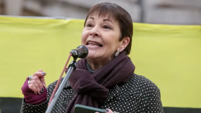 Caoline Lucas speaking at a microphone with a light green banner behind her. She is wearing a polka dot coat and has a dark purple scarf around her neck.