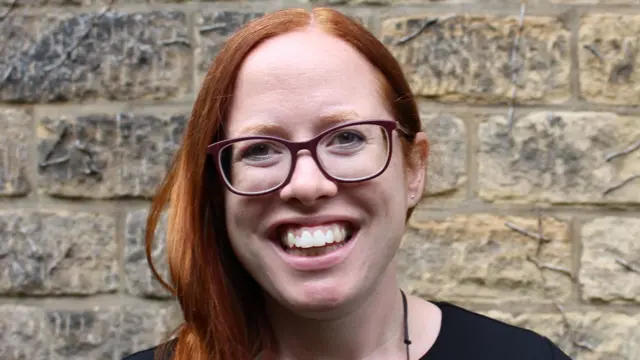 A woman with red hair and glasses stands smiling in front of a brick wall