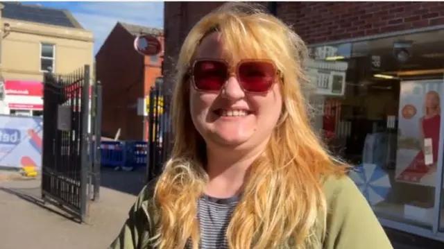 A woman smiles while wearing sunglasses in a town centre. She is wearing a green cardigan and striped t-shirt.