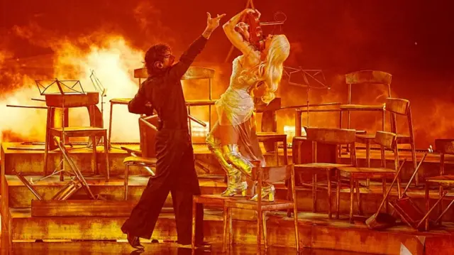 Finland's entry performing on stage, he's in a black suit and she is in a silver outfit with a violin