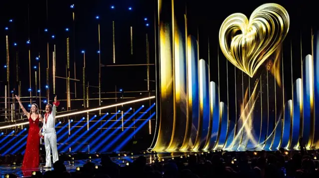 Eurovision stage 2026, a huge gold heart is at the back of the stage with the presenters waving at the crowd at the front