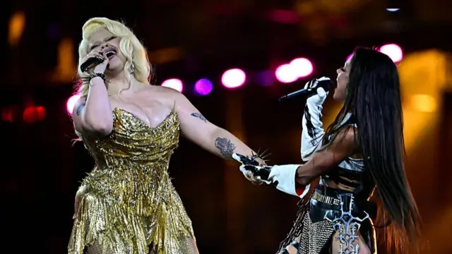 Conte in a gold dress and Ruslana in a black and white outfit embracing one arm while with their other hands they sing