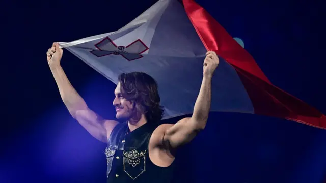 A man holding a Maltese flag and a cowboy vest smiling