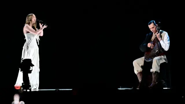 Victoria sings in a white dress against a black background. A man sits in front of her playing an instrument. he sits on a chair while she stands