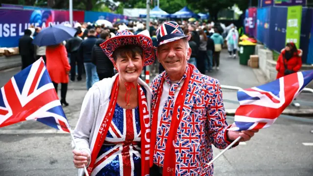 A man and woman hold one another as they smile and face the camera. They wave Union Jacks and wear Union Jack clothing and hold flags