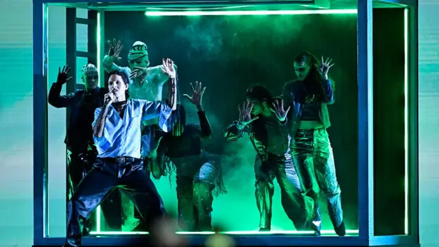Lund performing in front of five dancers in a big glass cube that is lit in green and there is smoke floating around the floor