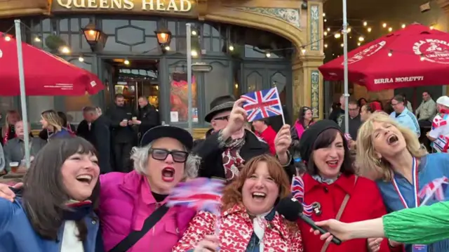 Fans at a pub waving British flags and screaming into a microphone while smiling and laughing