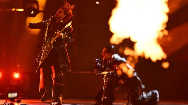 A man playing guitar while another kneels and screams into a mic. Flames are in front of them