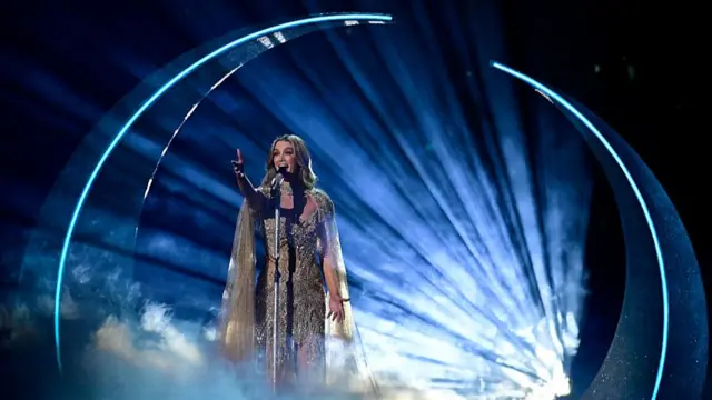 Delta singing in a gold gown in front of a crescent moon prop and a huge spotlight casting beams out behind her