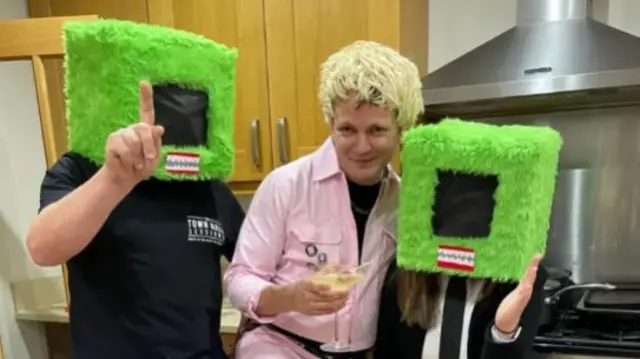 Jonathan in a blonde wig and pink outfit with two people wearing furry TV's as helmets