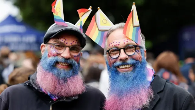 Two men pose together and smile looking forwards. They have dyed their large bears blue and pink, and have lgtbq+ flags sticking up from their ears and jewels on their face. They look very happy