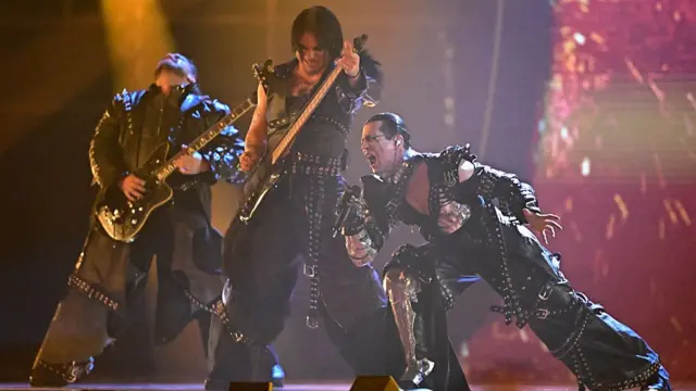 Three members of Lavina on stage singing and wearing huge black outfits with lots of chains and metal accessories