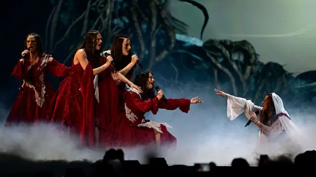 Four female singers on the left in large red dresses pointing at a singer on the right who kneels in a white dress and looks on at the women pointing at them. She is covered partially by smoke