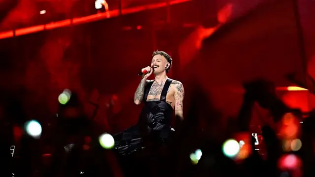 Lovv standing in dungarees on stage singing into a mic. The background is lit up red