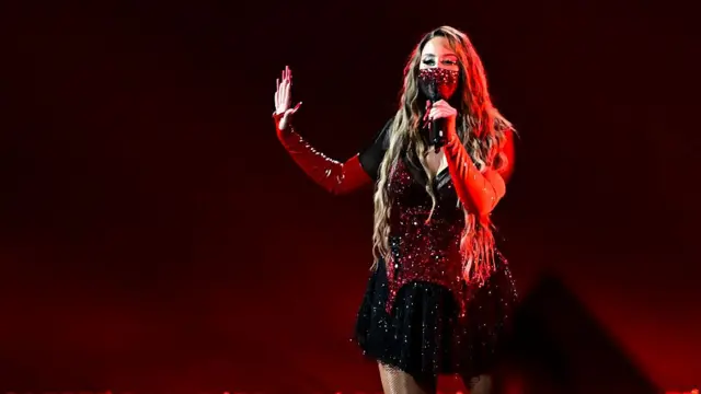 Felicia stands singing in a red and black sequined dress with one hand held out to the side and the other holding a mic. She has a face mask covering her mouth and nose