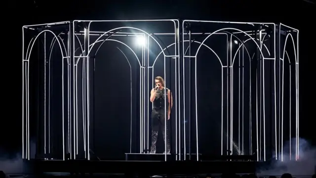 Aidan stands in a circle of archways in front of a black background