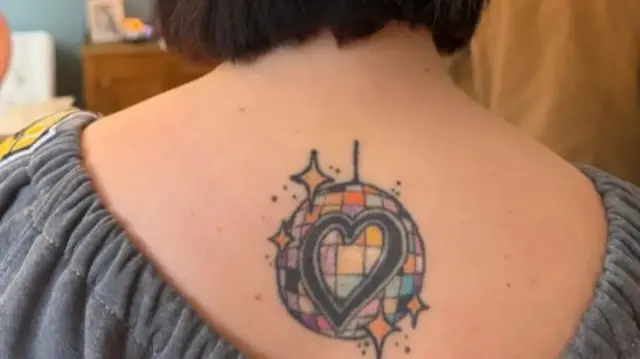 A picture of a colourful disco ball on someones back just below their neck. The disco ball is multi coloured and has a black heart in the middle of it
