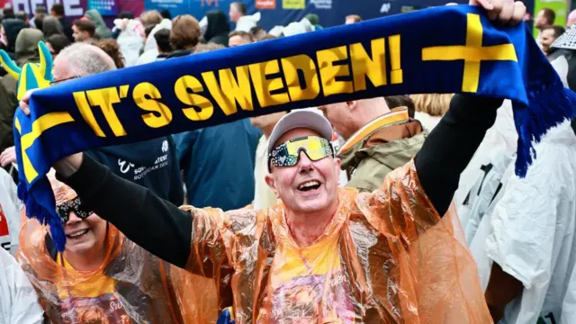 A man holding a sweden scarf up in a crowd wearing huge reflective yellow glasses