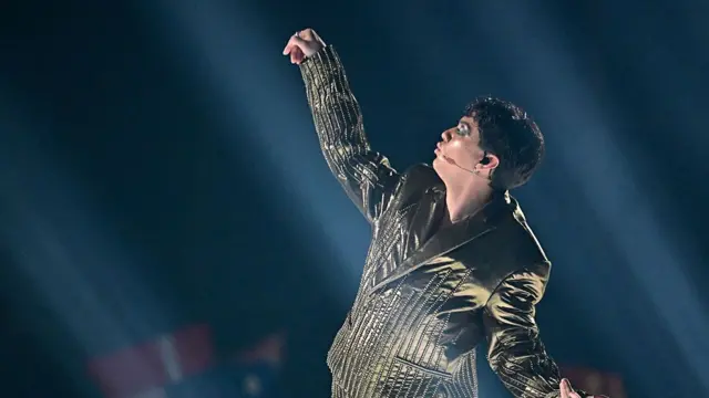 JJ stands with his arms apart looking up as he sings against a dark background