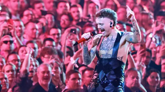 Lovv on stage singing into a microphone. He wears sparkly dungarees and has tattoos on his arm and chest. A crowd of people watch on behind him.