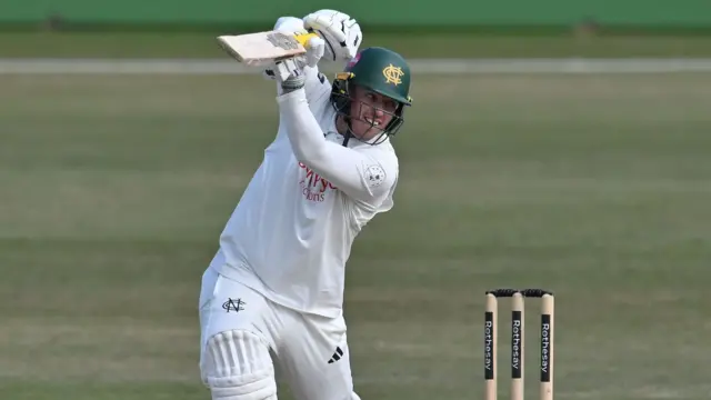 Ben Slater, wearing a green helmet, white pads, white gloves and holding a bat, plays a shot while batting for Nottinghamshire