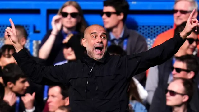 Pep Guardiola, in a black zip-up coat, holds his arms outstretched and shouts in front of Chelsea fans