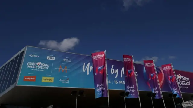 Eurovision flags flying outside a large arena against a blue sky with some clouds