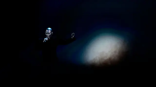 A silver man with a silver head singing into a mic holding his hands up in the darkness