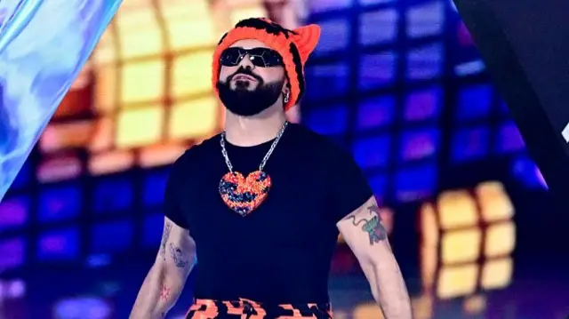 Akylas looks up wearing an orange beanie and large orange heart necklace and sunglasses