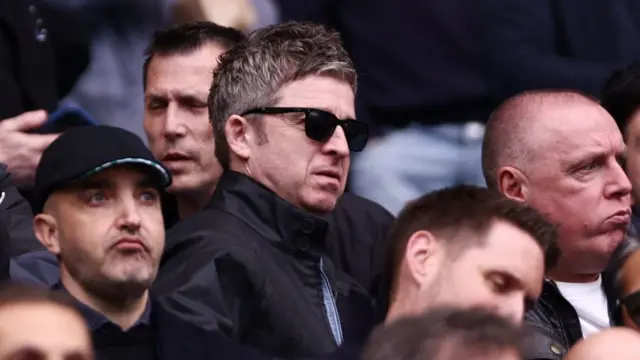 Noel Gallagher, wearing dark shades, among fans at a Manchester City game