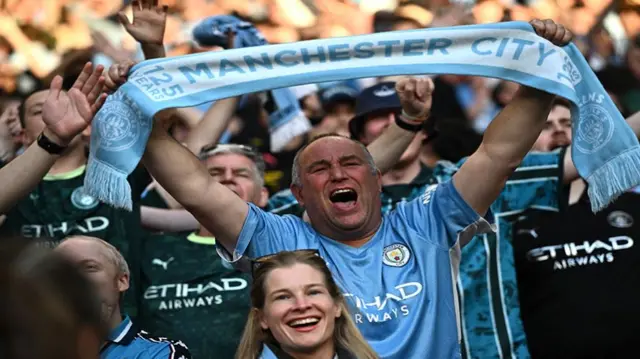 Manchester City fans in the crowd celebrating the FA Cup semi-final win with one holding up a club scarf
