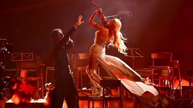 A man looking up and reaching his hand up at a woman who faces him on a chair playing a violin