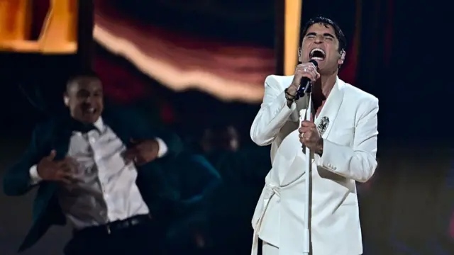 A man with a white suit emotionally singing into a microphone as a man dances behind him