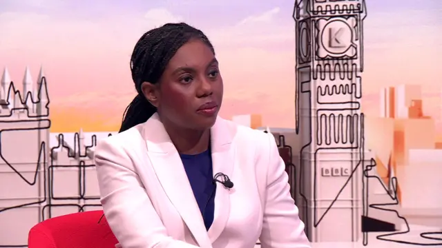 Kemi Badenoch in white blazer and blue dress sitting on a red chair