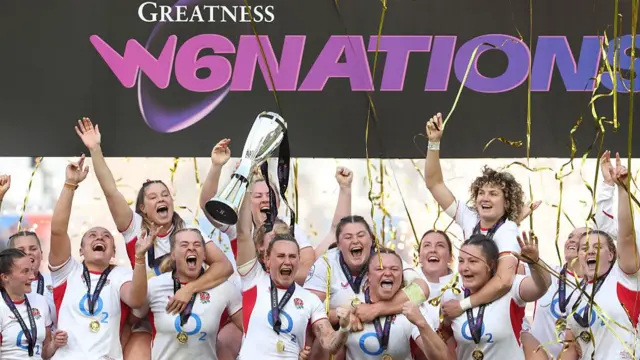 Meg Jones (centre) holds the silver trophy aloft in her right hand, surrounded by England team-mates (all wearing white England shirts with medals on blue ribbons around their necks)