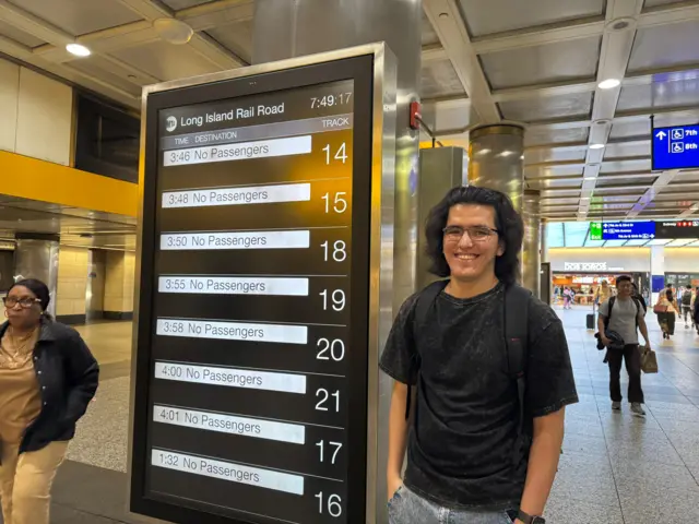 A man with long hair and glasses stands by a train departure board with no trains shown departing