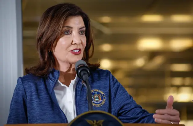 New York Governor Kathy Hochul in a blue zipped sweater and white shirt speaks into a microphone