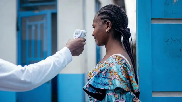 A woman in an African print dress has her temperature taken. A hand, holding an electronic thermometer, can be seen stretching out from the left of the picture.