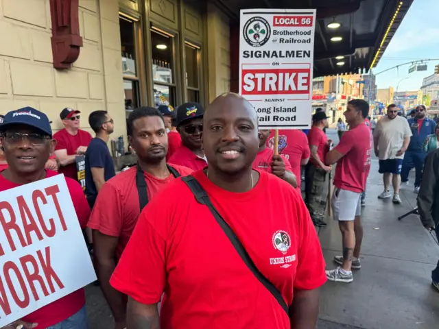 A man in a red t-shirt looks into the camera as picketers hold protest signs around him