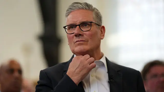 Prime Minister Keir Starmer pictured wearing white shirt and black suit jacket with dark-rimmed glasses. He appears to adjust collar, while other people can be seen faintly in the background
