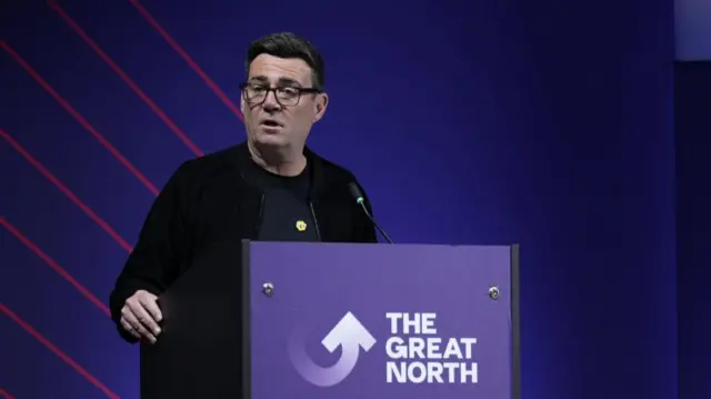 Andy Burnham in a black jacket and t-shirt addressing a conference while standing from behind a purple lectern, the front reading The Great North
