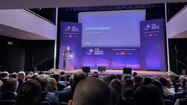 Wide shot of a conference hall, Andy Burnham speaking behind a purple lectern on the left-side of the stage