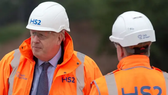 Boris Johnson wears an orange hi-viz jacket with 'HS2' written on it. The jacket is open and a blue shirt and tie can be seen. He is also wearing a hardhat that says 'HS2'.