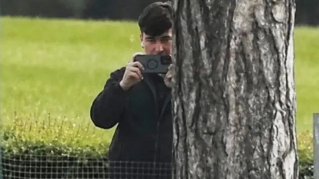 A man standing next to a tree holding a phone as if filming or taking a photo. He has short dark hair and is wearing a zip-up black top.