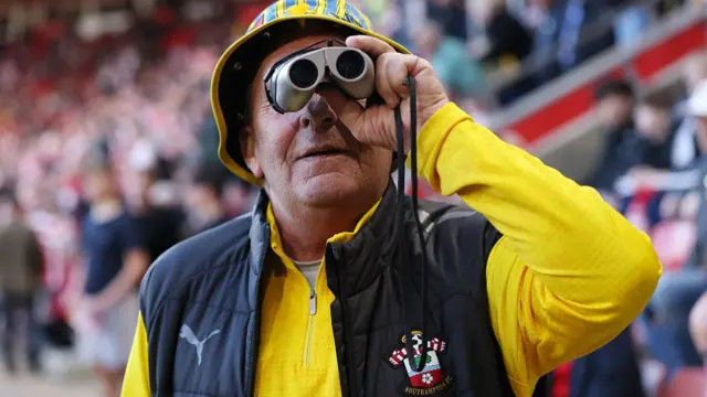 A man wearing a yellow Southampton shirt and a black gilet holding binoculars and a yellow hat.