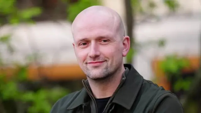 Stephen Flynn smiles. He is wearing a dark green jacket and appears to be standing in a garden.