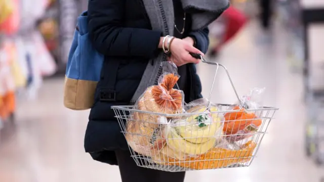 A woman holding a grocery basket that includes bread, bananas and carrots.
