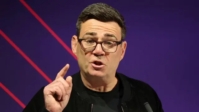 Greater Manchester Mayor Andy Burnham gesturing while speaking against a purple background.